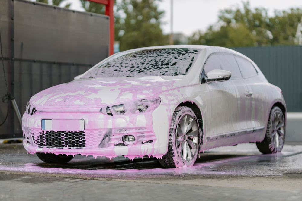White sedan covered in pink foam during a professional car deep cleaning service in Dubai. High-pressure contactless wash ensures a spotless, scratch-free finish.