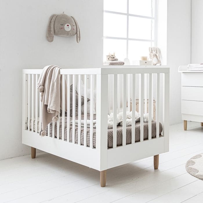 Minimalistic white baby crib with a beige blanket draped over it, featuring soft, neutral-toned bedding and a cute bunny wall decoration in a bright, airy nursery.