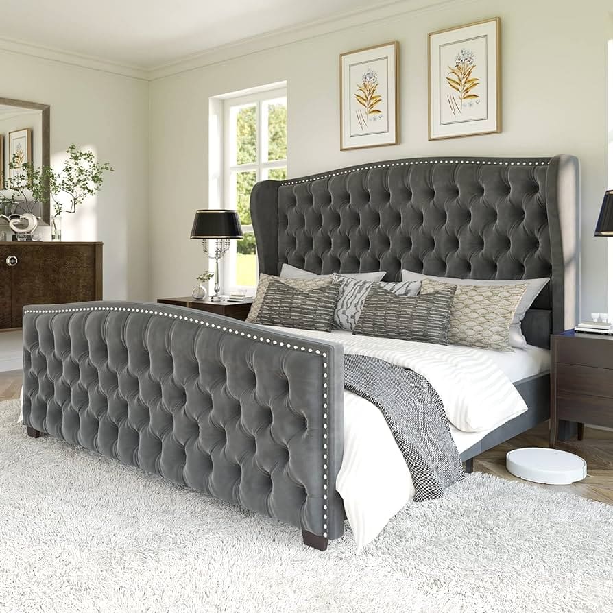 Elegant tufted gray velvet bed with a high headboard and matching footboard, adorned with decorative pillows and a cozy blanket, set in a light-filled bedroom with framed botanical artwork on the walls and wooden furniture.
